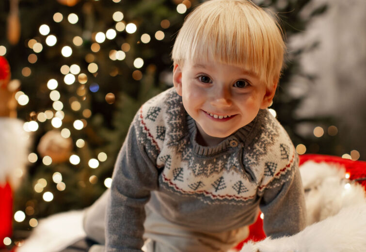 Uśmiechnięte dziecko na drewnianych saniach ozdobionych światełkami, z rozświetloną choinką w tle