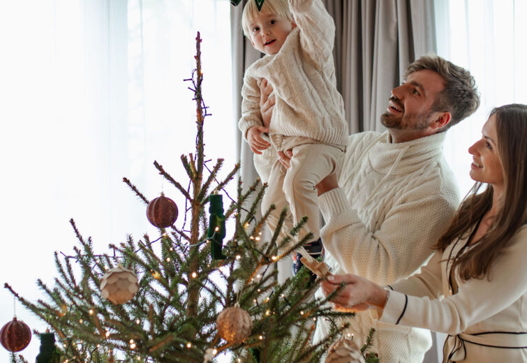 Rodzina dekorująca świąteczną choinkę w jasnym salonie z dużymi oknami i zasłonami.