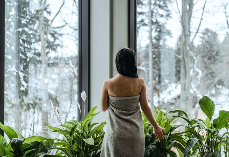 Kobieta owinięta ręcznikiem podziwia zimowy widok przez duże okna w relaksującym spa z zielonymi roślinami.