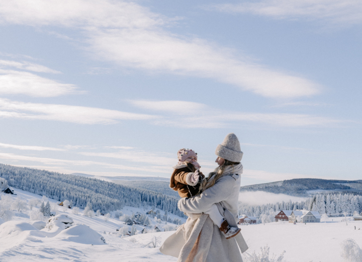 Woman in warm coat and hat holding child dressed for winter in snowy landscape with forested hills
