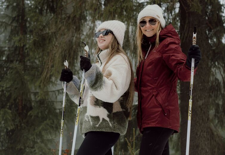 Two smiling women in warm jackets and hats cross-country skiing in a snowy evergreen forest.