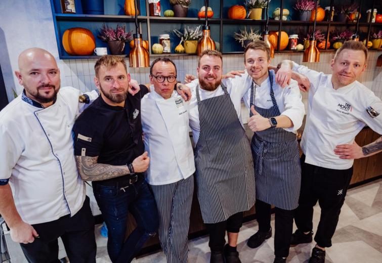 Group of six chefs in white and black aprons posing together in a restaurant kitchen with warm, inviting atmosphere