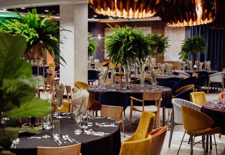 Elegant banquet room with round tables draped in black linens, lush fern centerpieces, and mixed colorful upholstered chairs.