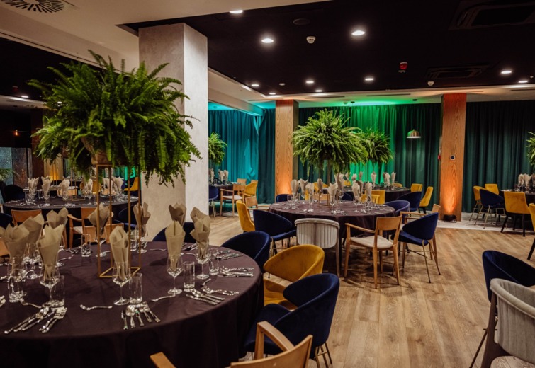 Elegant banquet hall with round tables, gold and navy chairs, and lush green plants as centerpieces for events.