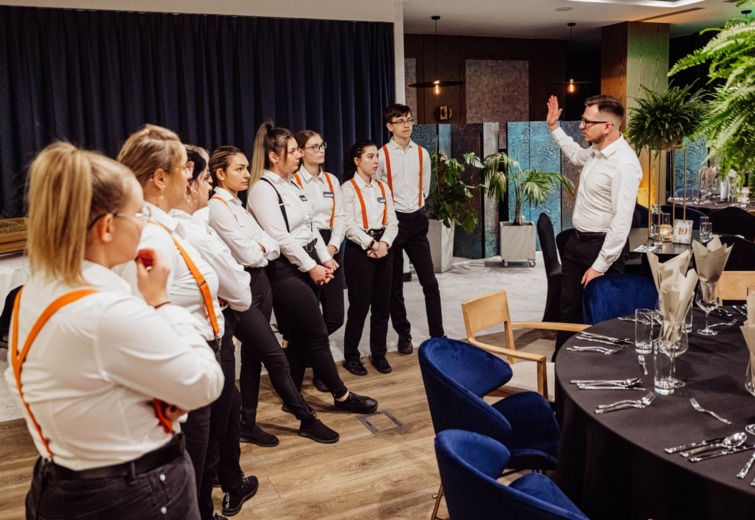 Hotel staff in white shirts and orange suspenders attending a training session in a modern banquet room with elegantly set tables.