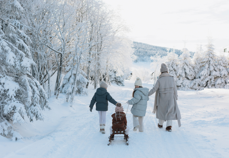 Family walking along a snowy winter path in a snow-covered forest, child sitting on sled pulled by an adult