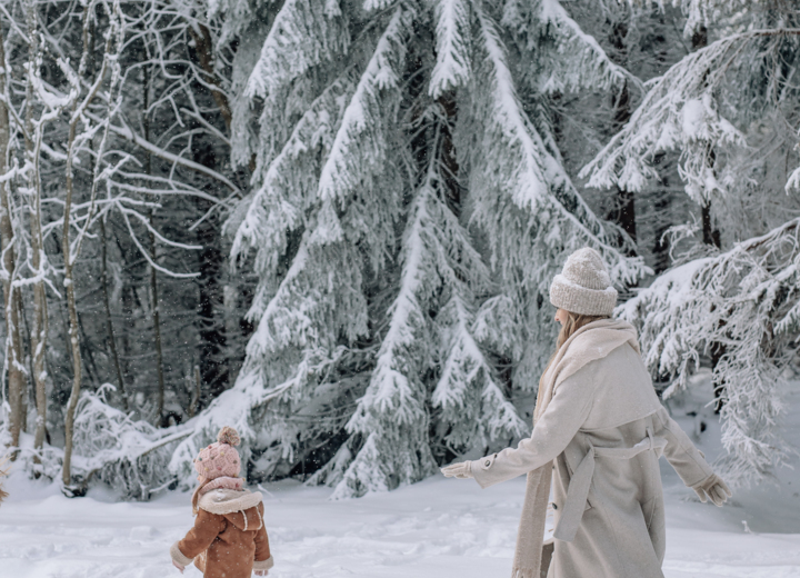Woman and child walk through a snowy forest path surrounded by snow-covered pine trees in winter.