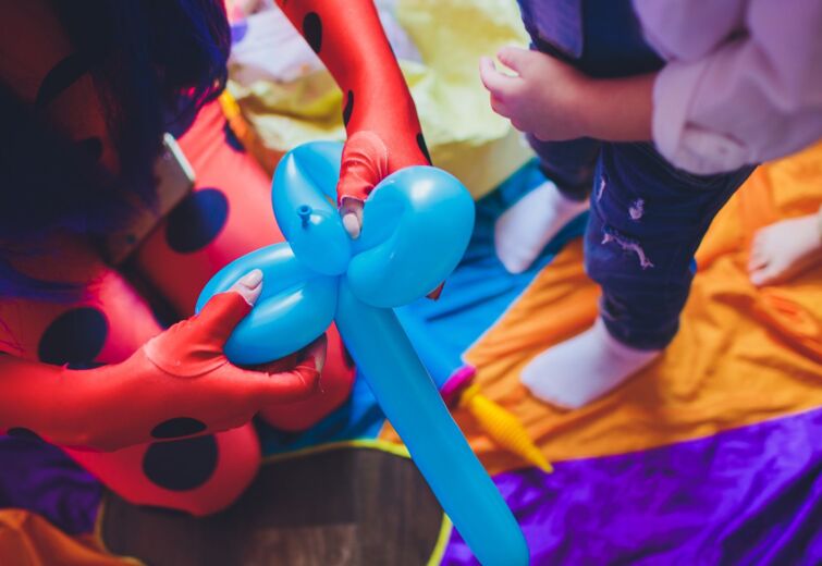 Animator wearing red gloves twists a blue balloon dog for children playing on a colorful mat