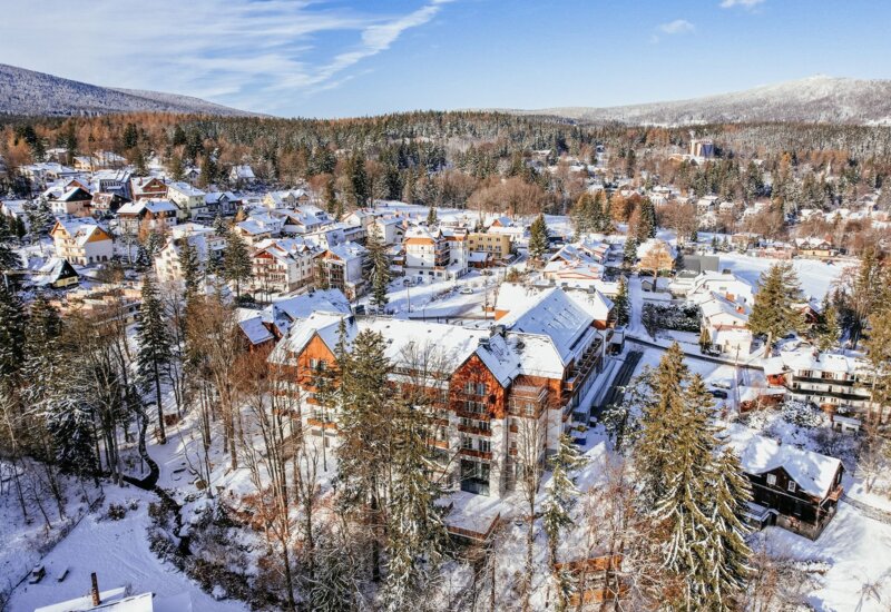 Zimowy widok na górski kurort z drewnianym hotelem, ośnieżonymi dachami i lasami w tle.