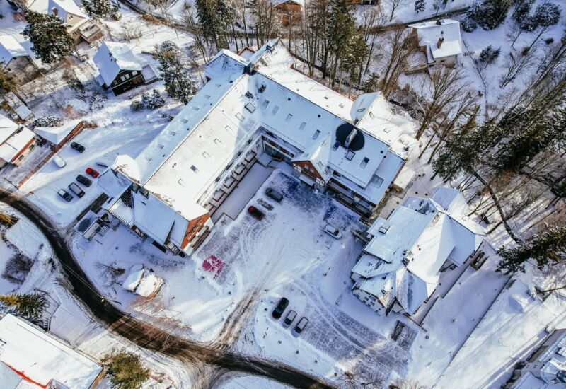 Hotel w zimowej scenerii z pokrytym śniegiem dachem, miejscem parkingowym i okolicznymi domkami w lesie