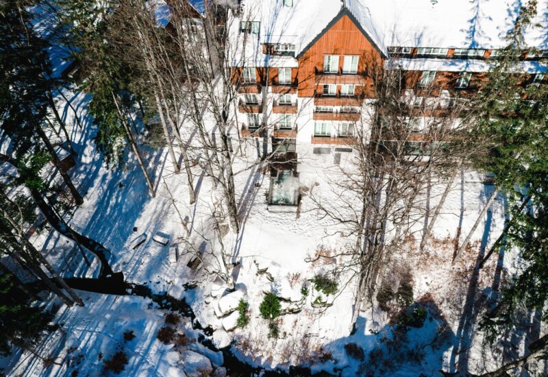 Górski hotel z drewnianą elewacją, ogrzewanym jacuzzi na zewnątrz i śnieżnym ogrodem otoczonym drzewami.