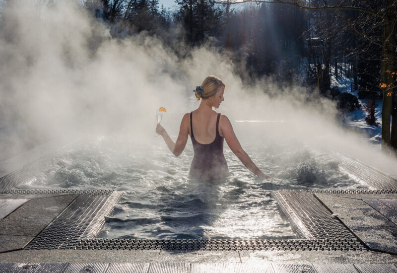 Kobieta relaksująca się w gorącej jacuzzi na zewnątrz otoczonej mgłą, trzyma kieliszek drinka, widok na zimowy las.