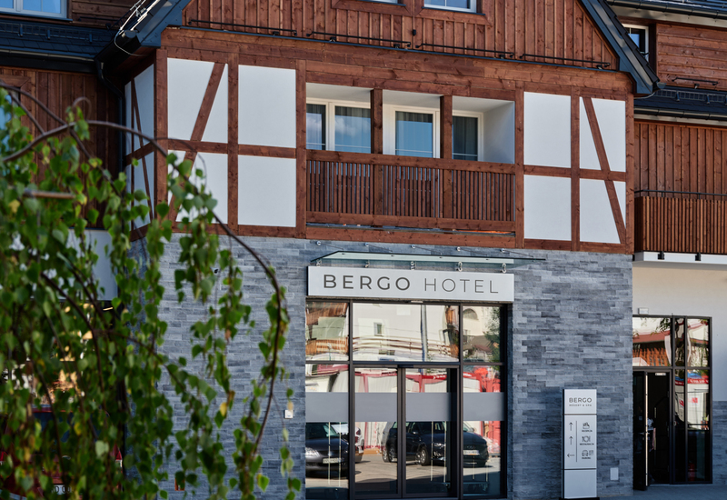 Wejście do hotelu Bergo z drewnianą elewacją i kamienną fasadą oraz balkonem nad wejściem.