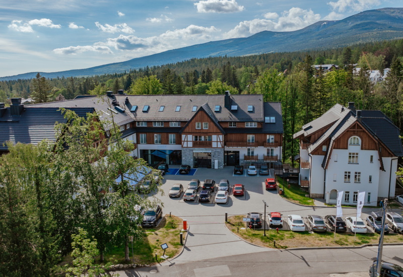 Hotel górski z tradycyjną drewnianą architekturą, parkingiem i widokiem na zalesione wzgórza i góry w tle.