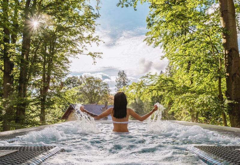 Gość relaksuje się w jacuzzi na świeżym powietrzu otoczonym bujną zielenią i promieniami słońca w rustykalnym otoczeniu.