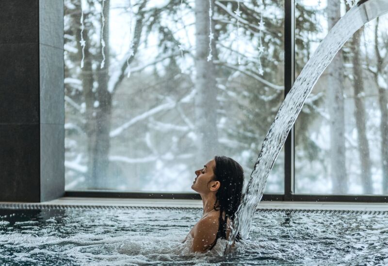 Gość relaksuje się pod strumieniem wody w krytym basenie z widokiem na ośnieżoną zimową scenerię za dużymi oknami