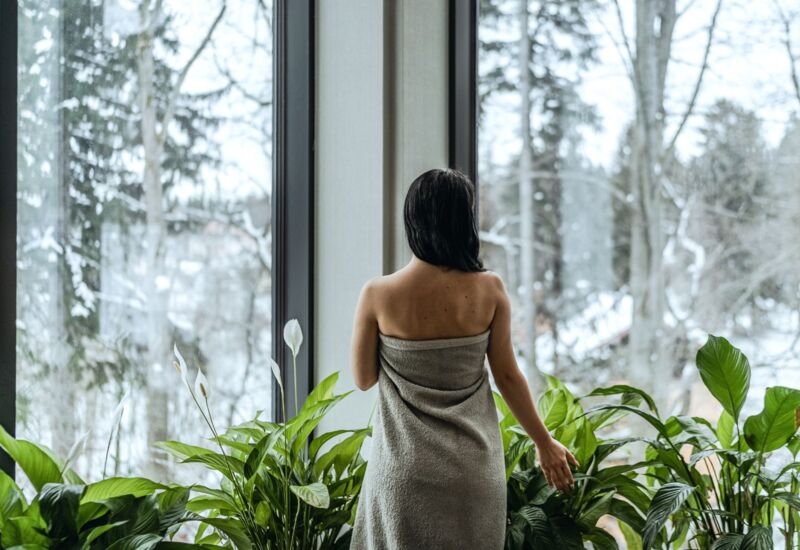 Kobieta owinięta ręcznikiem podziwia zimowy widok przez duże okna w relaksującym spa z zielonymi roślinami.