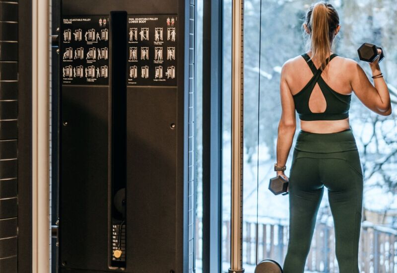 Gość w siłowni hotelowej ćwiczy z hantlami przy dużym oknie z widokiem na zimowy ogród na zewnątrz.