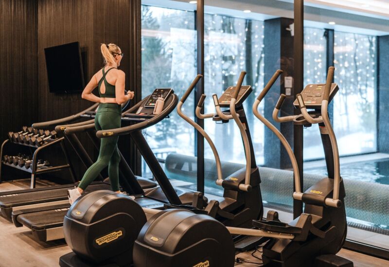 Kobieta biegająca na bieżni w nowoczesnej siłowni hotelowej z widokiem na basen przez duże okna.
