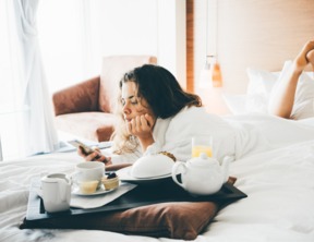 Gość w białym szlafroku odpoczywa na łóżku w jasnym pokoju hotelowym, delektując się śniadaniem z obsługi: czajnik, ciastka i sok.