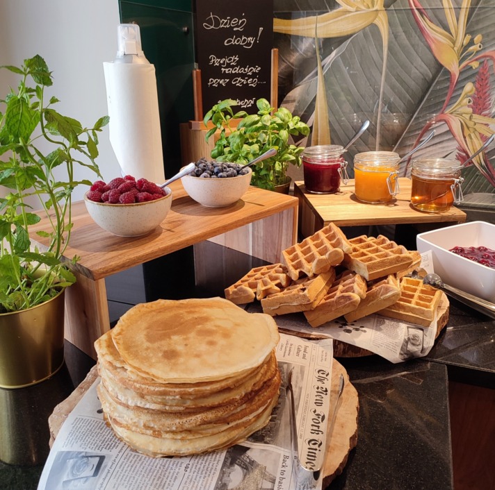 Stos naleśników i gofrów na hotelowym bufecie śniadaniowym, słoiki dżemów, miski owoców, świeża mięta i bazylia.