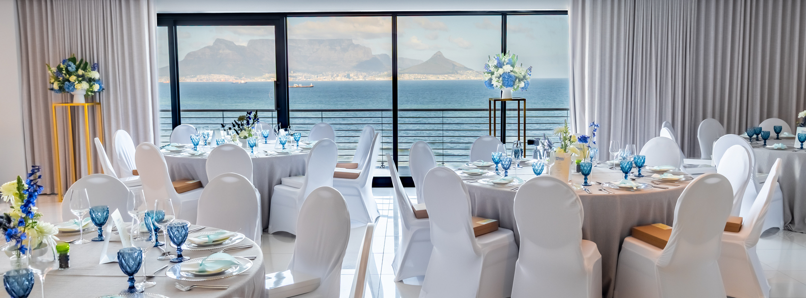 Ocean-view banquet room with round tables, white chair covers, blue glassware and floral centerpieces by floor-to-ceiling windows.