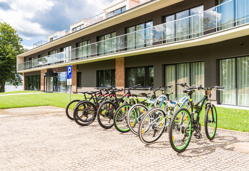 Fahrradverleih vor modernem Hotel mit Glasbalkonen, grünem Rasen und Blick auf das Wasser.
