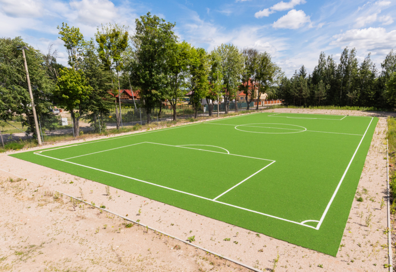 Zewnętrzne boisko sportowe z zieloną sztuczną trawą, otoczone drzewami, idealne do rekreacji na świeżym powietrzu