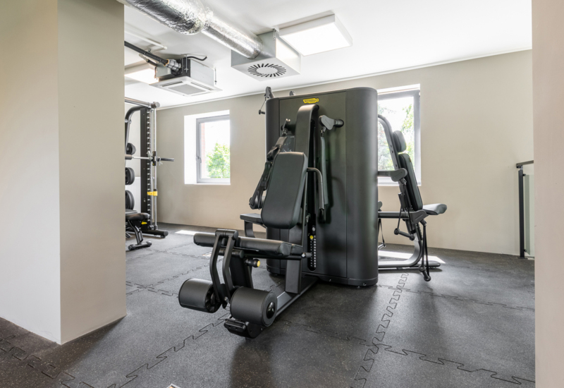 Nowoczesna siłownia hotelowa z różnorodnym sprzętem do ćwiczeń i naturalnym światłem przez okna.