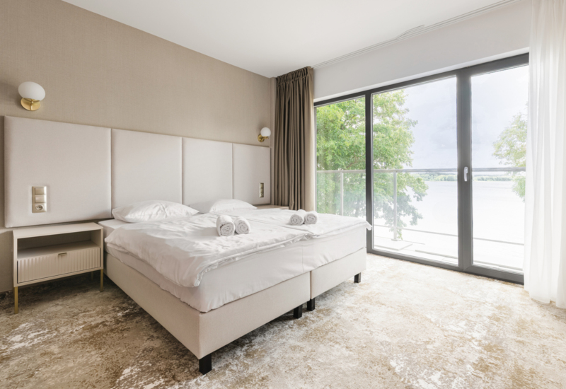 Jasny, nowoczesny pokój hotelowy z podwójnym łóżkiem, dużym oknem z widokiem na wodę i drzewami, dywanem i szafką nocną.