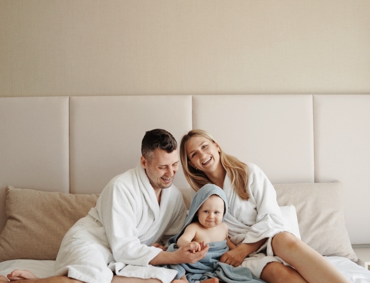 Familie entspannt sich auf einem großen Hotelbett in weichen weißen Bademänteln, Baby in Handtuch eingewickelt, gemütliche Atmosphäre