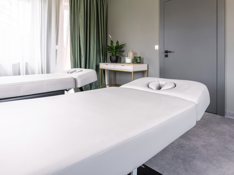 Nowoczesny pokój spa z dwoma białymi leżankami do masażu, zielone zasłony i stolik z rośliną i świecami