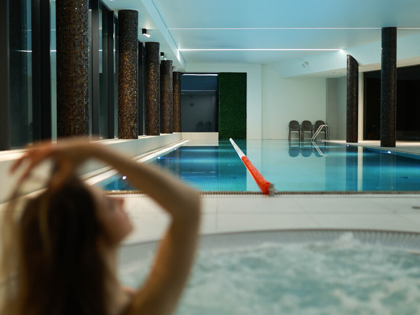 A person relaxes in a jacuzzi with a view of the indoor hotel pool. The calm atmosphere of the wellness area encourages rest and relaxation.