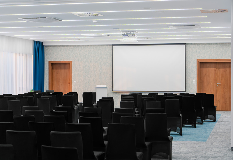 Sala konferencyjna z ustawionymi krzesłami, ekranem projekcyjnym i miejscem dla prelegenta. Przestrzeń podkreśla profesjonalny charakter spotkań nad morzem.