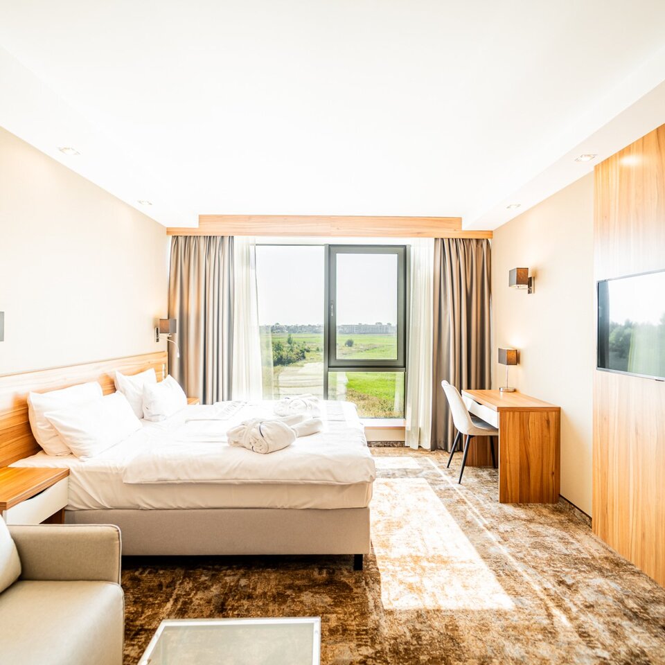 Ein helles Hotelzimmer mit großem Bett, Sofa, Schreibtisch und Fenster mit Blick ins Grüne. Das Interieur eignet sich ideal zum Entspannen.