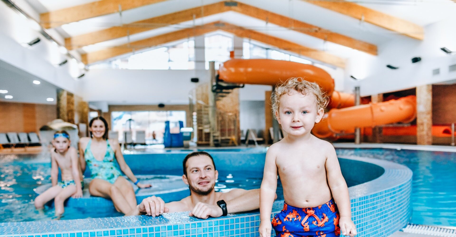 Family-friendly indoor pool with orange waterslide, mosaic-tiled edge and a smiling child in swim trunks in the foreground
