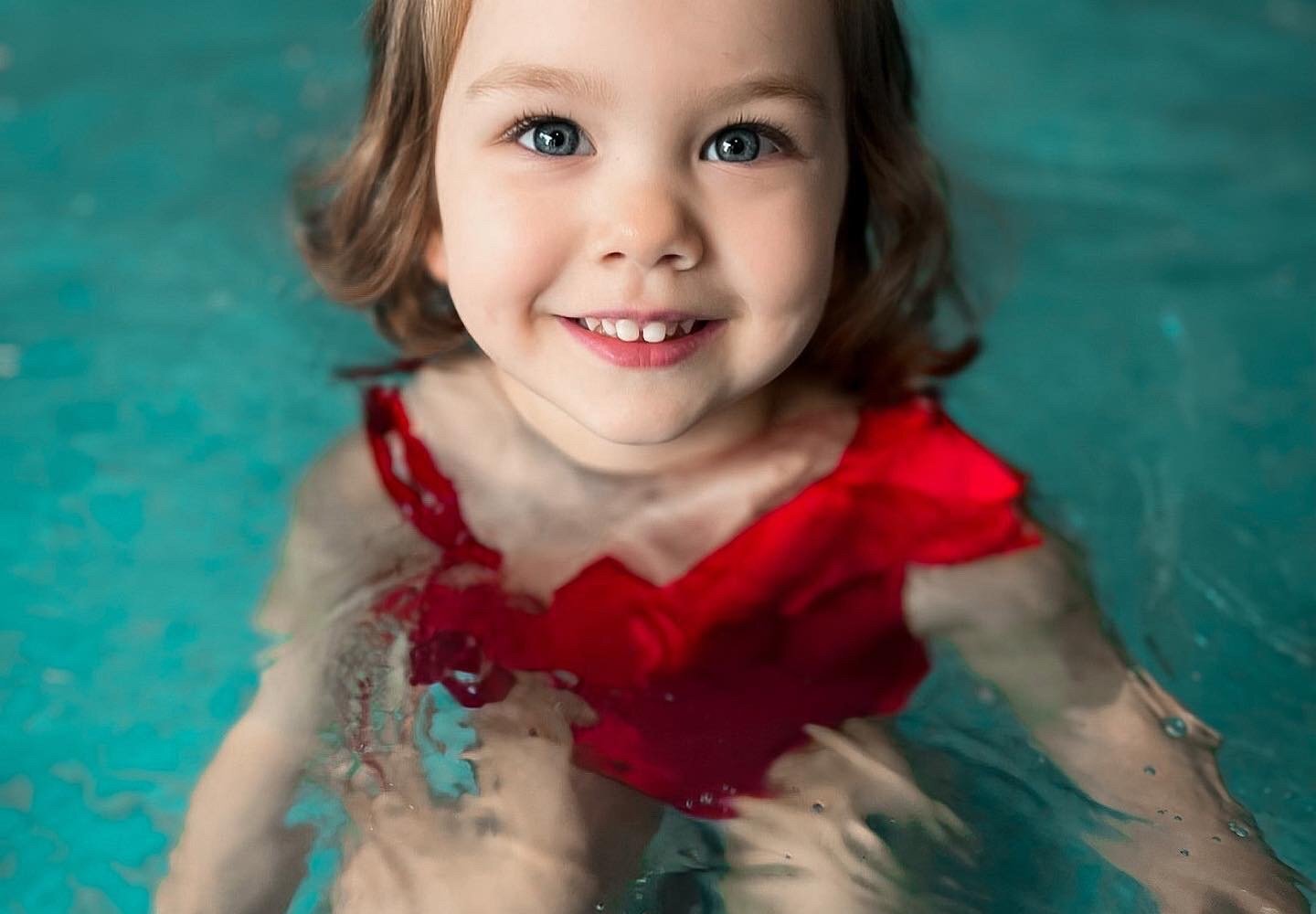Smiling young child in a red swimsuit standing in clear turquoise pool water