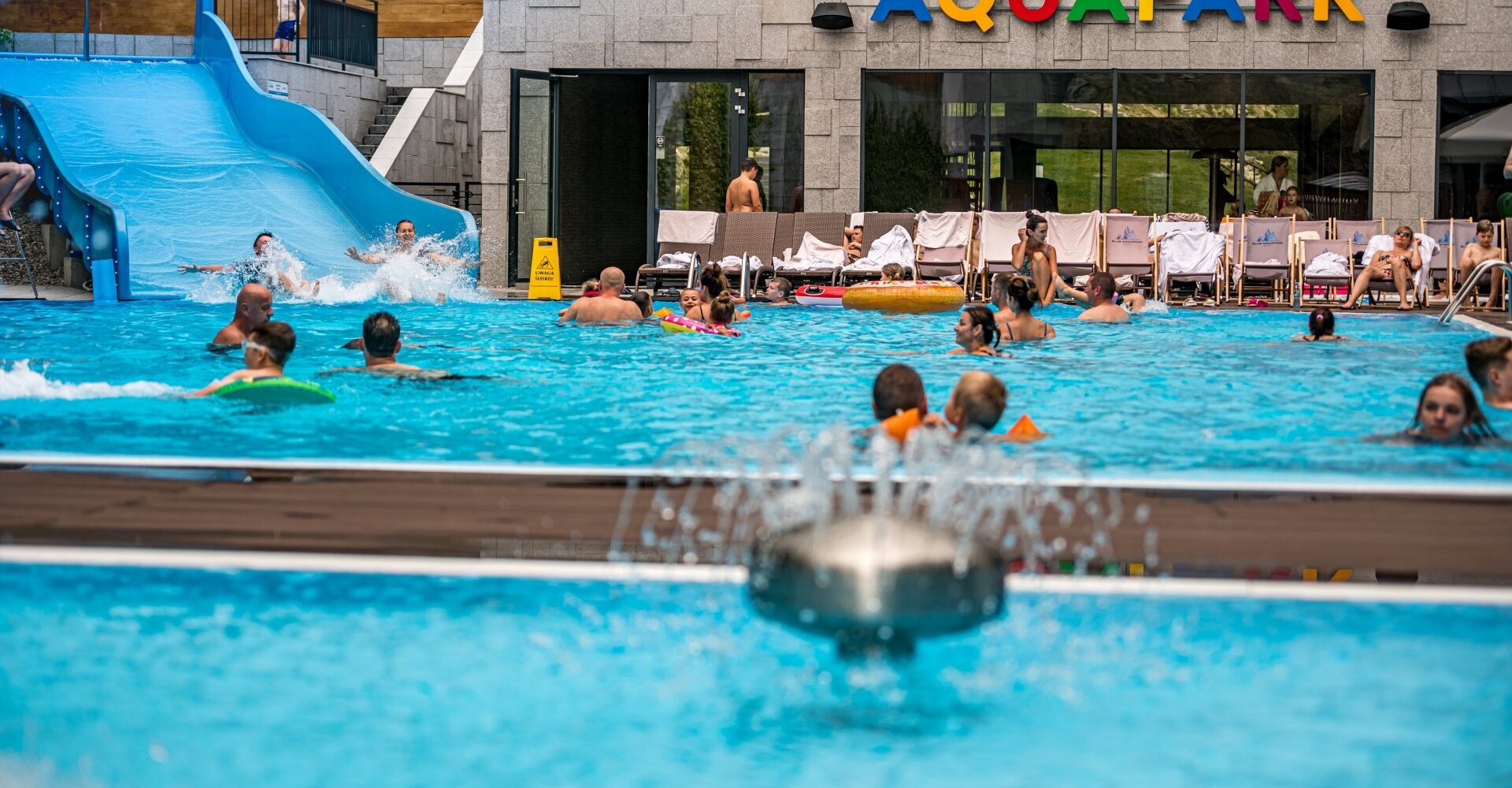 Outdoor aquapark with large blue water slide, families swimming, inflatable rings and sunloungers by a modern pool building.