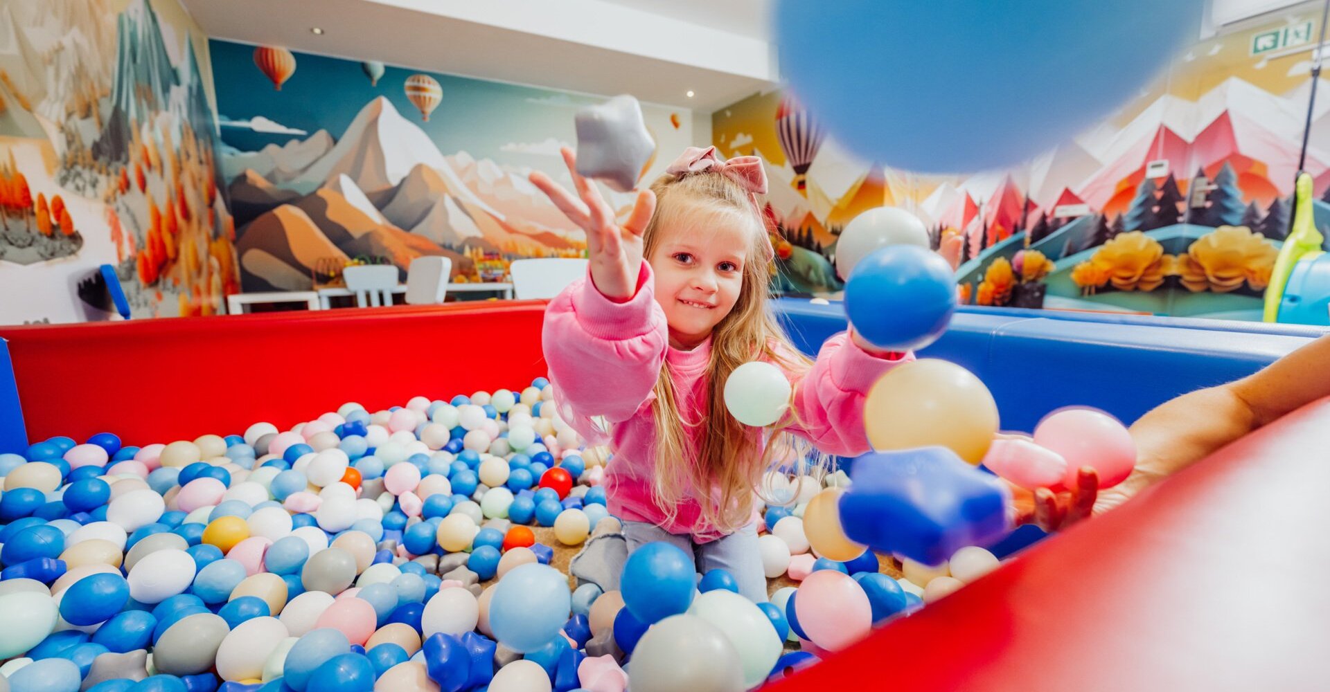 Jasna sala zabaw dla dzieci z basenem z kulkami i kolorowym muralem gór z balonami, przyjazna rodzinom.