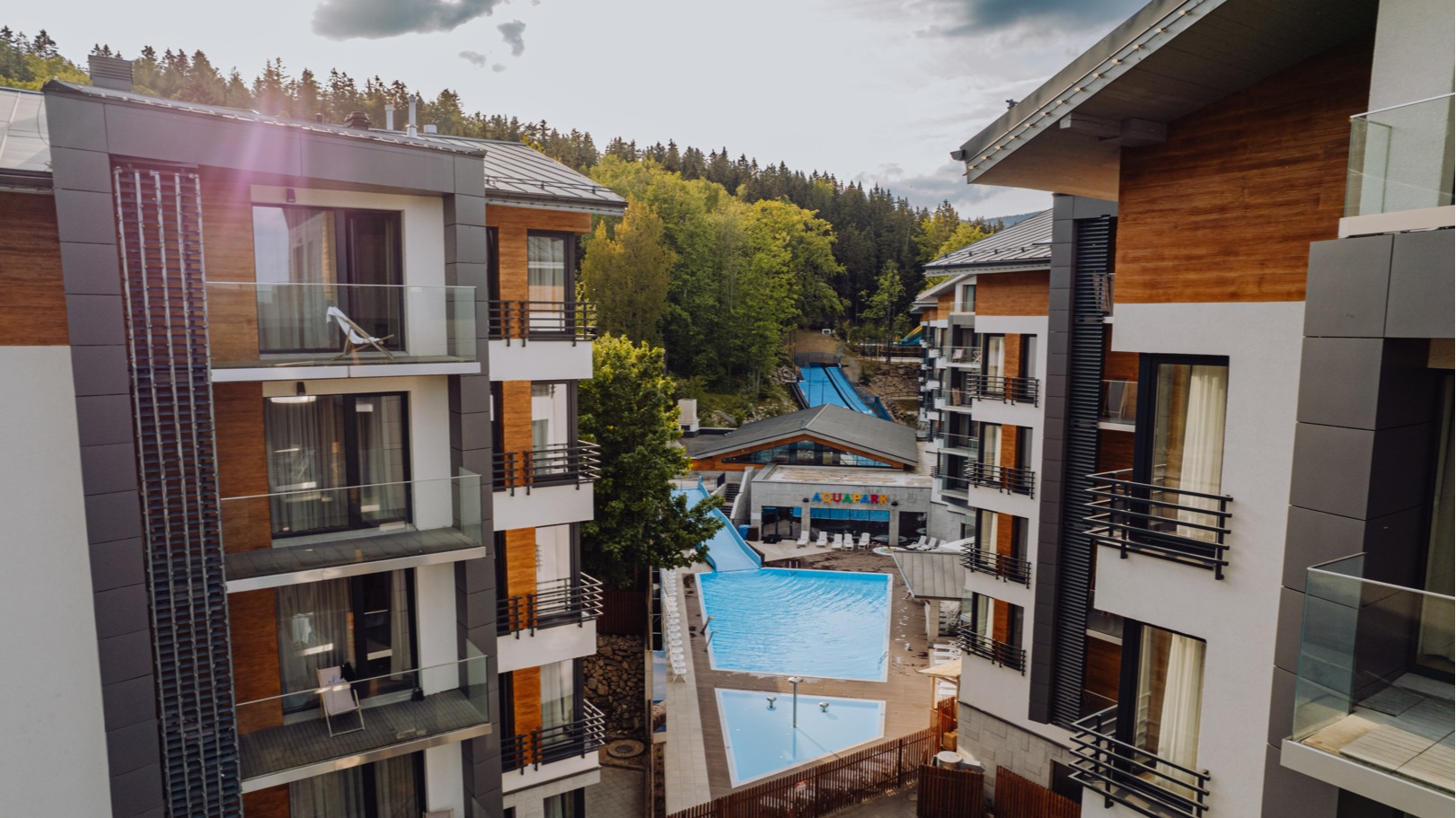 Modern mountain hotel complex with glass balconies, outdoor pool and aquapark slides set against a forested hillside.