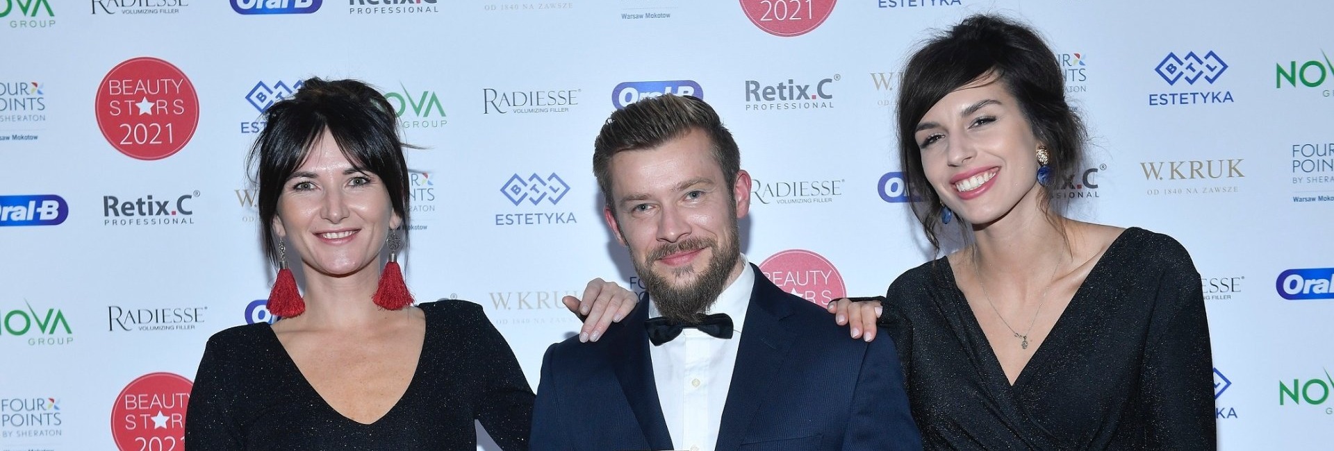 Trzy eleganckie osoby na gali, mężczyzna w smokingu trzyma statuetkę, dwie kobiety w czarnych sukienkach przed firmowym tłem.