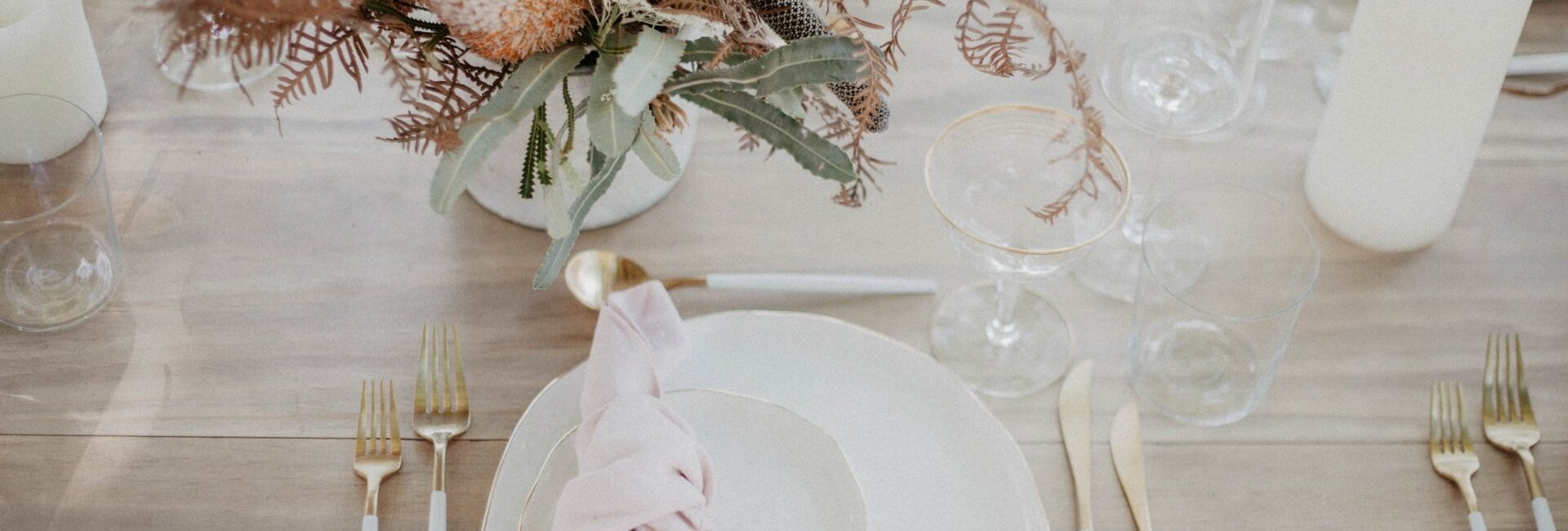 Elegantní svatební stůl s růžovou látkovou ubrouskou na bílých talířích, příbory se zlatými rukojeťmi, sklenicemi a sušenou květinovou dekorací