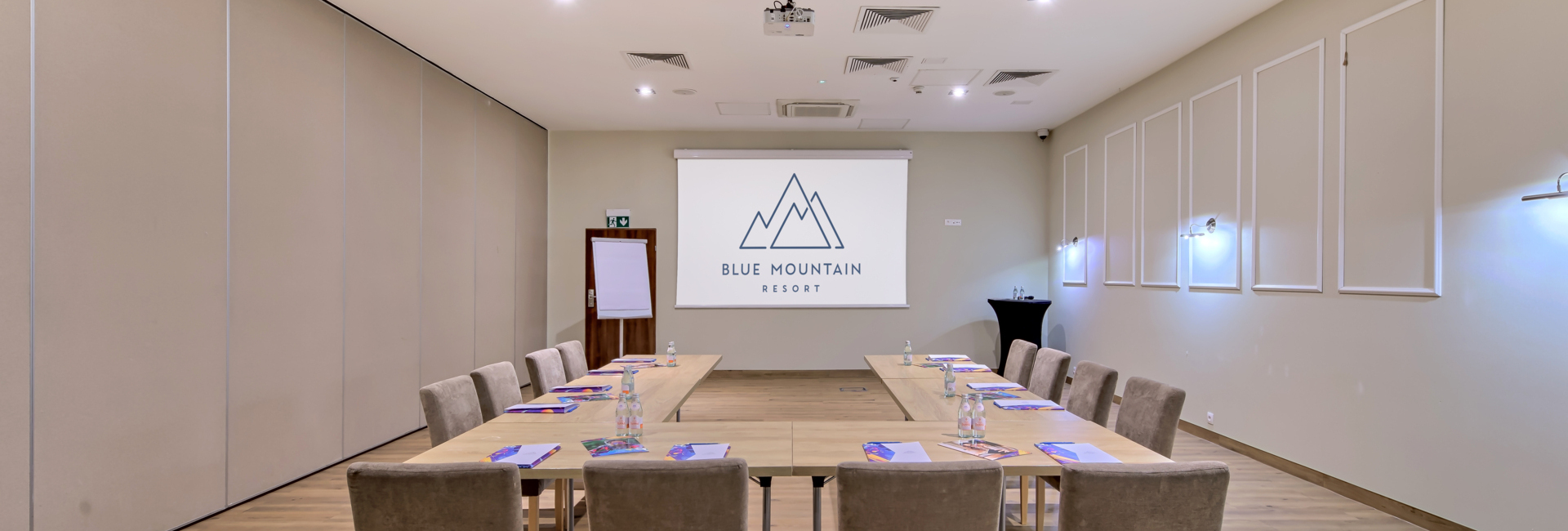 Konferenzraum in U-Form mit gepolsterten Stühlen, Projektorleinwand mit 'Blue Mountain Resort'-Logo, Flipchart und Wasser.