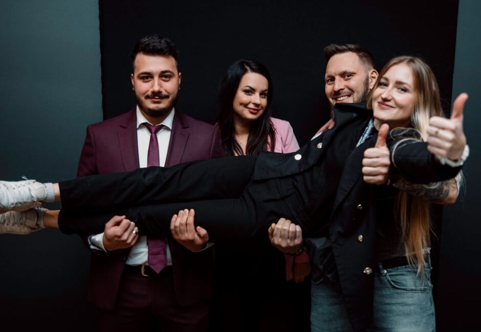 Usměvavý hotelový tým v oblecích a smart-casual drží kolegyni vodorovně, hravá atmosféra po akci
