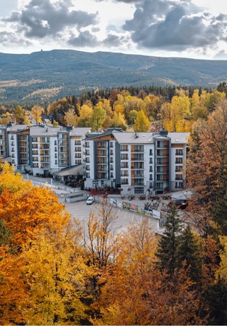 Moderní horský hotel s balkony v zlatém podzimním lese, parkovištěm a panoramatickým výhledem na hory.