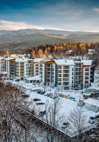 Modern mountain hotel with balconies, snow-covered grounds and parked cars, surrounded by forest and snowy peaks at sunrise