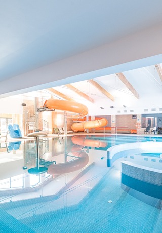 Indoor hotel pool with orange water slide, mosaic columns, lounge chairs and circular spa seating.