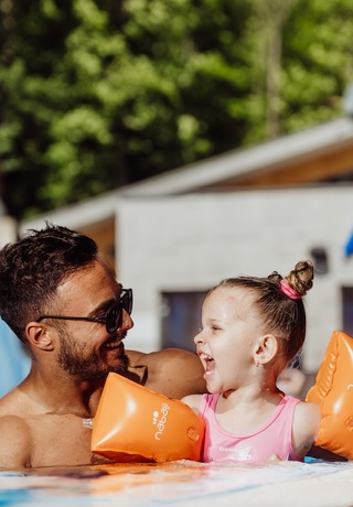 Father and young daughter laughing in outdoor hotel pool with orange arm floaties, pool ladder and colorful aquapark sign