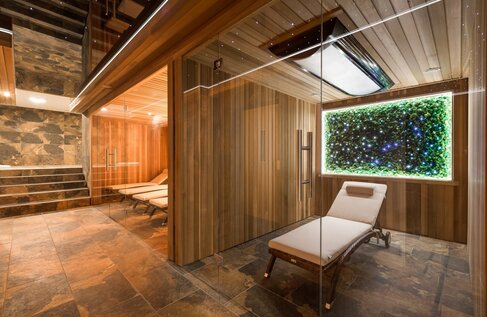 Wood-paneled hotel spa with glass-front sauna, heated lounge chairs, stone floor and illuminated moss wall.