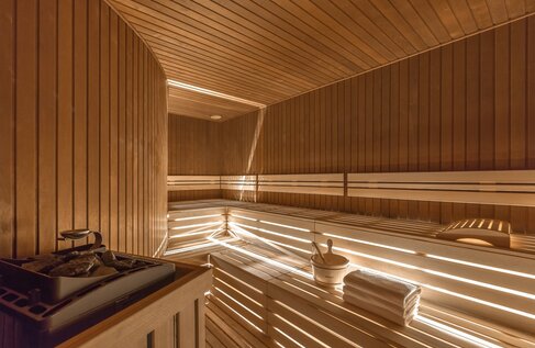 Wood‑panelled hotel sauna with tiered benches, warm recessed lighting, heater with stones, bucket and folded towels.